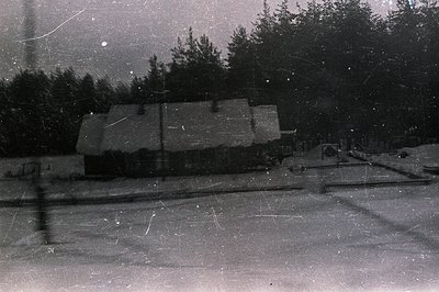 A snow-covered cottage with a steeply pitched roof nestles amongst dark evergreen trees. The monochrome image exhibits heavy ...