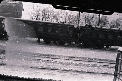A lengthy train with multiple passenger cars passes along parallel train tracks. The image is captured through a railing or g...