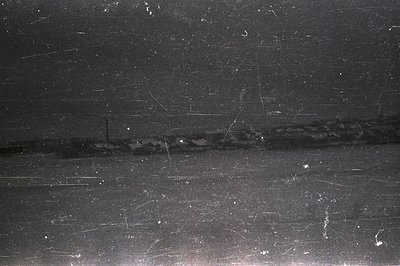 A heavily scratched black and white photo shows a coastal industrial landscape at dusk or night. A low-lying building complex...