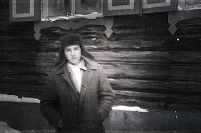 Man in a fur hat and coat stands before a log building. Visible signs of wear and scratches indicate age. Appears to be a por...