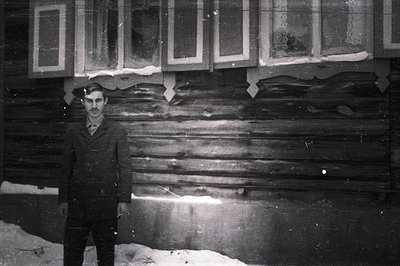 A man stands before a log building, likely a rural home. He wears a dark overcoat and tie, suggesting a formal or special occ...