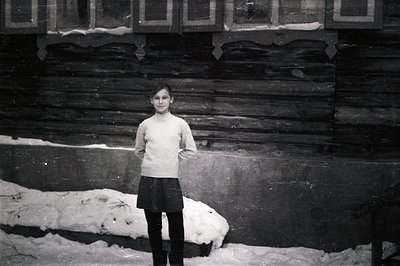 A young girl stands in snow before a timbered building, wearing a sweater, skirt, and dark stockings. The grainy black and wh...