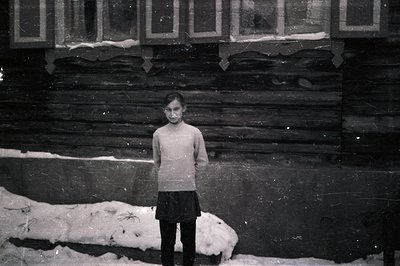 A young girl stands before a timbered building, snow visible at the base. She wears a simple sweater and skirt. The photograp...