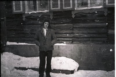 A young man stands in a snowy landscape before a rustic, log cabin-style building. He wears a textured overcoat and dark trou...