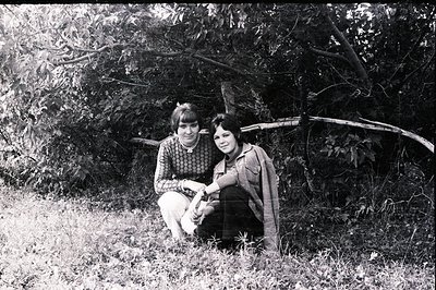 Two young women seated beneath a sprawling tree branch in a grassy, overgrown area. Both feature short, layered hairstyles ch...