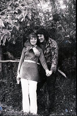A young couple stands embracing in a wooded area. The woman wears a patterned mini-dress and white bell-bottom trousers, whil...