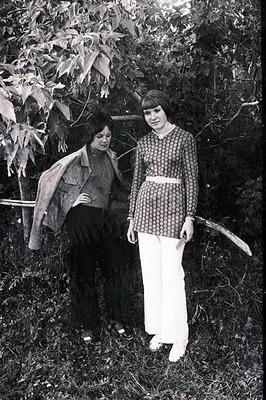 Two women pose outdoors amongst foliage. One wears a brown jacket, dark turtleneck, and black skirt. The other, in a patterne...