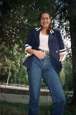 A young woman smiles confidently, styled in 1980s fashion: high-waisted jeans, white tee, and a navy blazer with white stripe...