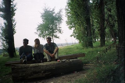Three young adults sit on a large fallen log amidst lush greenery, likely near a body of water. The photo's muted colors and ...