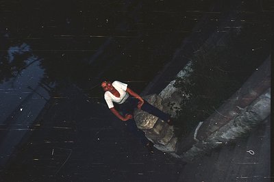 A young woman in a white t-shirt and jeans poses atop a large, weathered rock formation. The scene is shadowed, likely a grot...