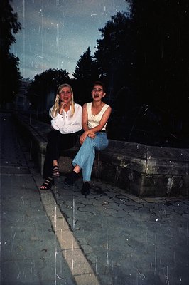 Two young women seated on a stone ledge, appearing relaxed and smiling. One wears a white blouse and dark sandals; the other ...
