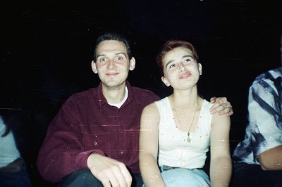 Two young adults, a man and a woman, pose closely, exhibiting relaxed smiles. The man wears a red-brown plaid shirt, while th...