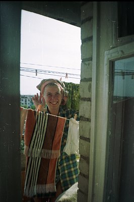 Man smiles from a doorway, wearing a plaid shirt, striped scarf, and head covering. Visible architecture suggests a seaside t...
