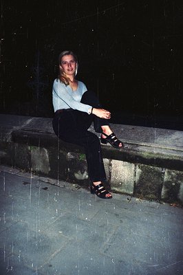 A young woman seated on a stone ledge, rendered in a grainy, low-light photograph. She wears a light grey sweater, black pant...