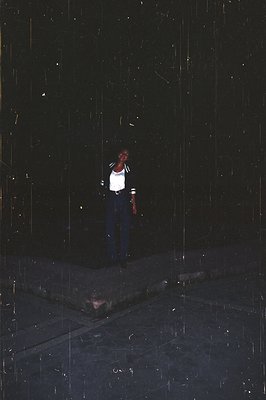 A young person stands on a rain-slicked street. They wear a white collared shirt, dark jacket with stripe detail, and blue je...