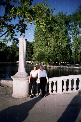 Two young women stand near a stone balustrade and column overlooking a river or lake, framed by lush green trees. They wear 1...