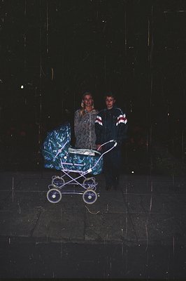 A young couple stands in the foreground, alongside a baby stroller with a patterned cover. They are dressed in casual 1990s a...