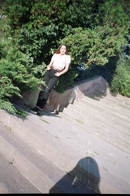 A young woman in a cream-colored top and dark jeans leans against a concrete hillside structure covered in dense greenery. Vi...