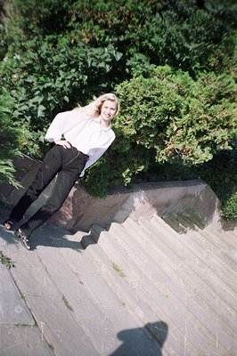 A stylish woman in a high-necked blouse and wide-leg black trousers poses on tiered concrete steps. Bright blonde hair and a ...