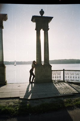 A lone figure stands silhouetted before a classical stone structure & ornate railing, overlooking a body of water. The photog...