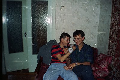 Two young men, likely friends, share a cup of tea in a cozy, floral-wallpapered room. Note the distinct 1980s fashion: stripe...
