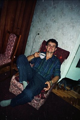 A man in dark plaid pajama set sits in a patterned armchair, holding a cup. Visible damage, including scratches and dust, imp...