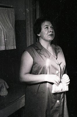 A woman in a button-down dress holds a clutch purse, gazing upwards. Likely a portrait from the 1950s or 60s, showcasing mode...