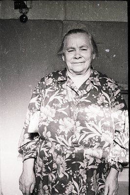 A candid portrait of an elderly woman with a gentle smile, standing against a textured wall. She wears a floral print shirt o...