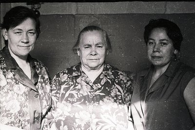 Three women pose for a studio portrait; the central woman is older, with a solemn expression. All wear patterned dresses, typ...
