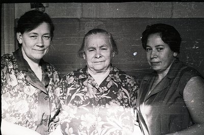 Three women stand closely together, facing the camera. The central figure appears older, with a calm expression. Both flankin...