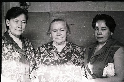 Three women stand closely together in a posed portrait, likely a family gathering. The central woman appears older, with a fl...