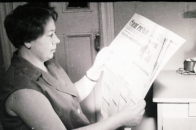 A woman reads a stack of newspapers, likely *Právo*, in a simple, functional interior. The scene, captured in black and white...