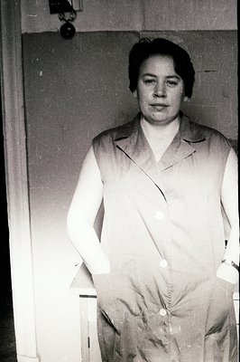 A woman stands formally in a button-up lab coat, hands clasped in front. Close-cropped dark hair and a serious expression cha...