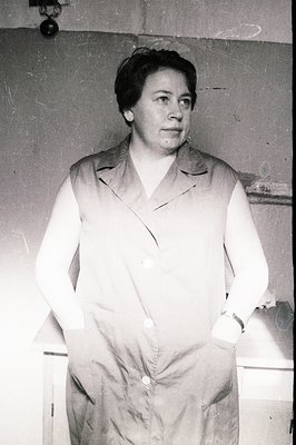 A woman in a functional, button-down work dress stands with hands clasped, facing the camera. The image, likely from the 1960...