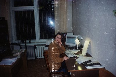 A young woman sits at a desk with a vintage computer setup. The room features a large window with a protective screen, sugges...