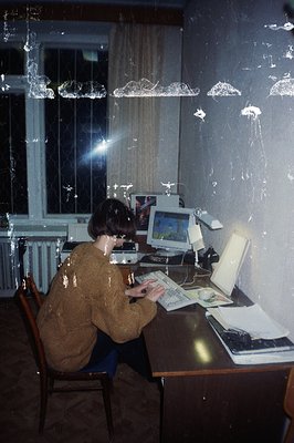 A person sits at a dark desk using an early personal computer, likely in the 1980s. The room's decor features patterned wallp...