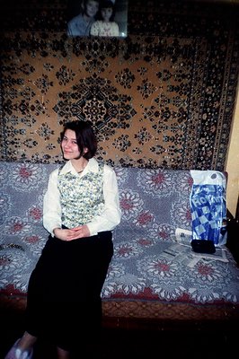 Young woman with bobbed hair, seated on a patterned sofa. She wears a white blouse, dark skirt and patterned vest. A framed p...