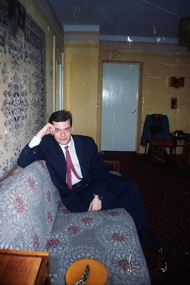 A man in a dark suit and red tie sits casually on a patterned sofa, hand resting on his knee. Interior view shows yellow wall...