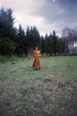 A woman stands in a field, framed by a dense evergreen forest under an overcast sky. She wears a long, brown wool coat and da...