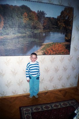A young boy stands in a room with a large landscape photograph on the wall. He wears a striped sweater and turquoise pants. T...