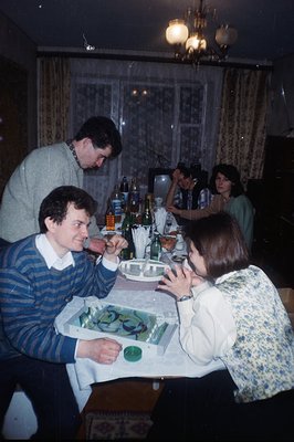 A family gathers for a meal, seated around a table laden with food and beverages. The decor suggests an interior from the 197...