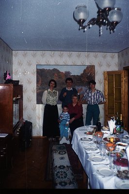 A formal dining room scene with a long table set for a gathering. Four figures – a woman in a patterned top, a young boy, a m...