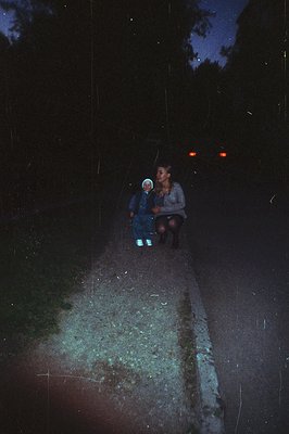 A mother and young child walk along a rural road at night. Visible headlights suggest an approaching vehicle, a common moment...