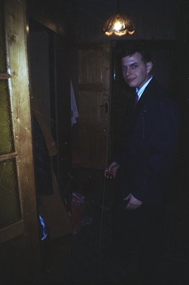 A young man in a dark suit and tie stands in a dimly lit hallway, posed near a doorway. Visible details include worn wooden p...