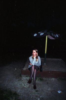 A woman sits on stone steps beneath a public shelter during rain. She wears a gray sweater, tights, and dark shoes. The scene...