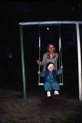 A young child in teal overalls and a matching hat sits on a swing held by a woman with shoulder-length hair. The scene is out...