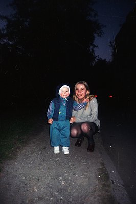A young child in blue overalls and a matching hat stands on a gravel path, accompanied by an adult woman squatting beside the...