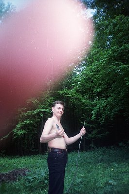 A shirtless man stands in a grassy area with lush green foliage, holding a thin, flexible branch. Distinctive 1970s style: hi...