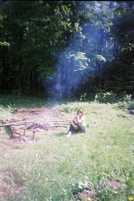 A lone figure sits near a small fire pit within a dense, green wooded area. Smoke rises from the fire. Appears to be a candid...