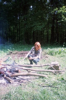 A young woman with red hair sits near a recently extinguished campfire in a grassy clearing surrounded by dense trees. She’s ...